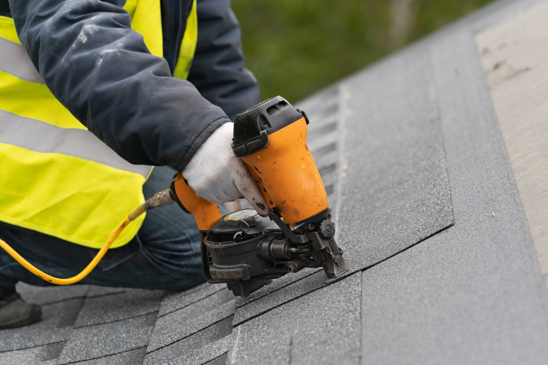 Qualified roofer worker in protective uniform using air or pneumatic nail gun and installing asphalt or bitumen shingle on top of the new roof under construction residential building